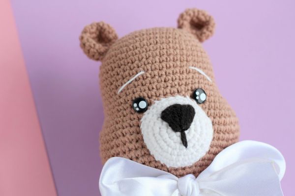 Close-up of a handmade crochet teddy bear with a white bow tie, set against pastel background.